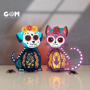 May include: Two decorative Día de Muertos paper lanterns, one a dog and the other a cat. The dog is blue with a floral crown, and the cat is purple with a rose. Each holds a glowing lantern with a cutout design. The lanterns are decorated with sugar skull designs.