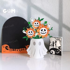 Puede incluir: Un arreglo artesanal de papel con temática de Halloween con un jarrón blanco en forma de fantasma que sostiene flores naranjas y blancas con caras de fantasmas. También hay una tarjeta negra en forma de lápida con la palabra "Halloween".