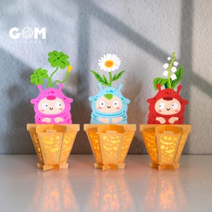 Può includere: Tre vasi di fiori di carta colorati con personaggi dei cartoni animati. Ogni vaso è di un colore diverso: rosa, blu e rosso. Ogni personaggio ha un fiore diverso in cima. I vasi hanno un design decorativo e sono illuminati dall'interno.