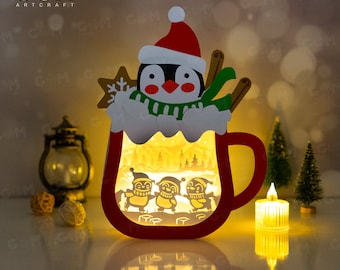 May include: A red and white penguin wearing a green scarf and a Santa hat is peeking out of a red and white mug. The mug has a window that shows three penguins ice skating in a snowy forest. The mug is lit from within, creating a warm glow.