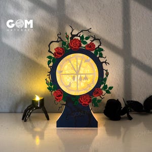 May include: A decorative, laser-cut wooden lamp with a circular design, featuring red roses and a gothic theme. The lamp has a dark blue base and frame, with a warm yellow light emanating from within. A small candle holder is in the foreground.