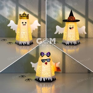 Puede incluir: Tres linternas de papel iluminadas con forma de fantasma y patrones recortados intrincados. Un fantasma lleva una corona dorada, otro un sombrero de bruja negro con flores naranjas, y el tercero gafas de sol, un lazo morado y un bolso naranja.