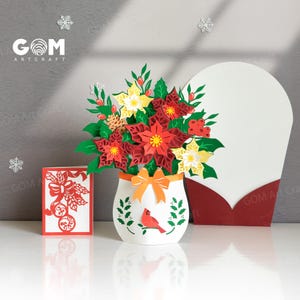 Op de afbeelding: Een 3D-papierkunst kerstarrangement met rode en gele poinsettia's in een witte vaas met een rode kardinaal. Een oranje strik siert de vaas. Een rode en witte kaart en een grote witte kaart zijn ook aanwezig.