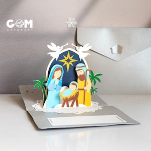 Pu&ograve; includere: Un biglietto di Natale pop-up con una scena della Nativit&agrave;. Il biglietto presenta Maria, Giuseppe e Ges&ugrave; bambino, realizzati in carta azzurra, gialla e marrone. Lo sfondo &egrave; blu scuro con una stella dorata. Il biglietto &egrave; aperto, rivelando la scena 3D.