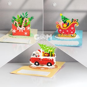 Pode incluir: Três cartões pop-up com tema natalino. Um apresenta um envelope vermelho com bengalas doces e vegetação. Outro mostra um trenó vermelho com presentes e uma árvore de Natal. O terceiro mostra um caminhão vermelho com o Papai Noel e presentes.