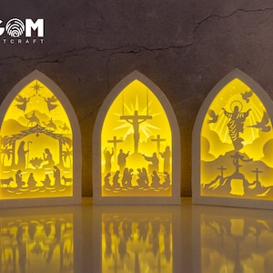 Puede incluir: Tres escenas iluminadas recortadas en papel que representan la Natividad, la Crucifixión y la Resurrección de Jesucristo. Cada escena está enmarcada en un arco blanco y se coloca sobre un fondo oscuro.
