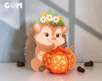 May include: A whimsical papercraft hedgehog with a floral crown and a smiling face, holding an orange pumpkin lantern. The lantern has a cut-out design and is lit from within. The image includes the text "GOM Artcraft".