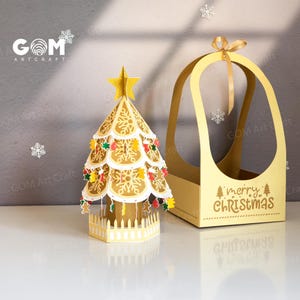 Peut inclure: Un sapin de Noël doré découpé au laser, surmonté d'une étoile et orné de décorations. À côté, un coffret cadeau doré assorti avec un ruban et l'inscription "merry christmas". L'arbre est sur une surface blanche.