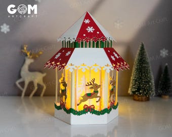 May include: A decorative paper lantern in the shape of a carousel, featuring a red and white roof with snowflake cutouts. The lantern has reindeer and Christmas tree cutouts, illuminated by a warm light. The brand name "GOM Art Craft" is visible.