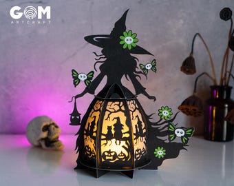 May include: A black papercraft witch with a green hat and a glowing lantern. The lantern has a black silhouette of a witch and a cat inside. The witch is holding a lantern and a broom. The lantern is lit up with a warm yellow light. The witch is surrounded by green leaves and flowers.