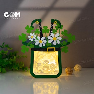 Puede incluir: Una lámpara de papel recortada verde y blanca con forma de sombrero de duende con tréboles y flores. La lámpara está encendida y muestra una escena de tres duendes dentro del sombrero.