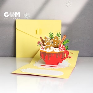 Puede incluir: Tarjeta de Navidad desplegable con una taza roja llena de galletas de jengibre, bastones de caramelo y palitos de canela. La tarjeta es amarilla con un sobre a juego. El texto "GOM Art Craft" es visible.