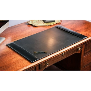 Könnte beinhalten: Ein schwarzer Schreibtischunterlage aus Leder mit erhöhten Kanten auf einem Holzschreibtisch. Ein Stift mit grünem und goldenem Design liegt auf der Unterlage. Der Schreibtisch hat Schubladen mit Messingbeschlägen. Das Holz hat einen warmen, rötlich-braunen Ton.