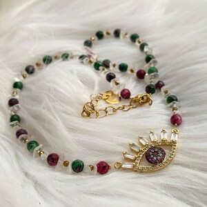 Könnte beinhalten: Eine goldene Halskette mit einem großen bösen Augen-Anhänger. Der Anhänger ist aus Gold gefertigt und hat ein blaues Auge mit einer roten Pupille. Die Halskette besteht aus weißen, grünen und roten Perlen.