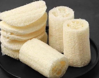 May include: A collection of natural loofah sponges in various shapes. Some are cylindrical, while others are sliced into flat, round pads. The sponges are a light beige color and have a porous, fibrous texture. They are arranged on a black surface.