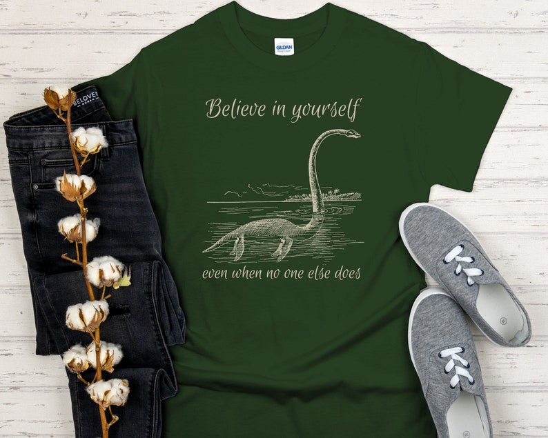 May include: A dark green t-shirt with a white graphic of a sea monster and the text "Believe in yourself even when no one else does".