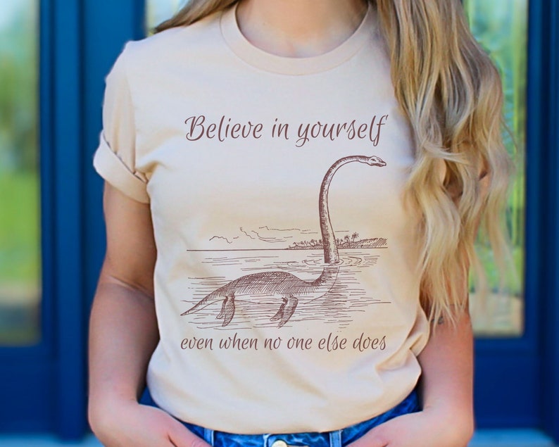 May include: A beige t-shirt with a brown line drawing of a plesiosaur with the text "Believe in yourself even when no one else does."