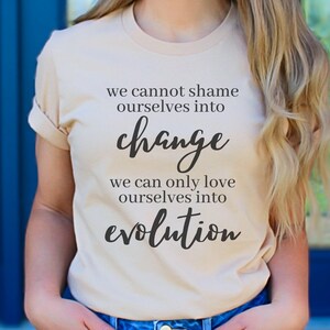 Puede incluir: Una camiseta beige con el texto "We cannot shame ourselves into change. We can only love ourselves into evolution." impreso en negro.