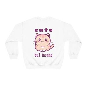 Może przedstawiać: Biała bluza z kresk&oacute;wkowym kotem i napisem "cute but insane" w kolorze fioletowym.