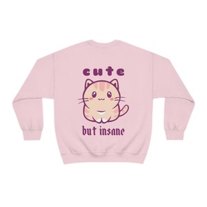 Może przedstawiać: R&oacute;żowa bluza z kresk&oacute;wkowym kotem i napisem "cute but insane" w kolorze fioletowym.