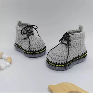 Könnte beinhalten: Ein Paar hellgraue gehäkelte Stiefeletten mit schwarzen Schnürsenkeln, einer dunkelgrauen Sohle und einem dünnen gelben Akzentstreifen entlang der Basis. Die Stiefeletten haben ein hohes Design mit einer kleinen Zuglasche an der Ferse.