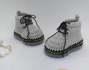 May include: A pair of light gray crocheted booties featuring black laces, a dark gray sole, and a thin yellow accent stripe running along the base of the upper. The booties have a high-top design with a small pull tab at the back of each heel.
