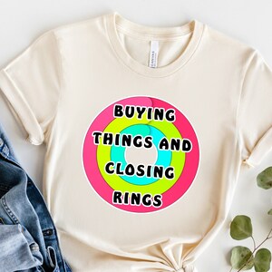 May include: A beige t-shirt with a colorful graphic design that says "Buying Things and Closing Rings".