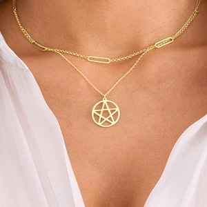 May include: A gold-coloured, layered necklace. The top layer has a chain with elongated oval links. The lower layer features a pendant with a circular frame and a five-pointed star design. The necklace is worn against a white shirt.