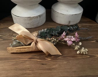 以下が含まれることがあります： ベージュのサテンリボンで結ばれたセージとパロサントの束。束には、乾燥したユーカリ、ピンクの花、小さな白い花が含まれています。アイテムは木製の表面に配置されています。