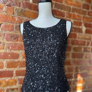 K&ouml;nnte beinhalten: Schwarzes Pailletten-Tanktop auf einer Schaufensterpuppe. Das &auml;rmellose Oberteil ist mit kleinen, gl&auml;nzenden schwarzen Pailletten bedeckt. Es hat einen runden Ausschnitt und eine taillierte Form. Der Hintergrund ist eine Backsteinmauer.