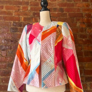May include: A colorful, open-front jacket with a unique patchwork design. The jacket features a mix of pink, red, orange, and blue geometric patterns. The sleeves are long and the jacket is displayed on a mannequin.
