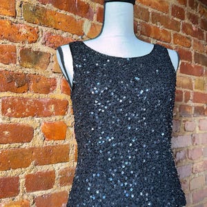 K&ouml;nnte beinhalten: Ein schwarzes Pailletten-Tanktop auf einer Schaufensterpuppe. Das &auml;rmellose Oberteil ist mit kleinen, gl&auml;nzenden Pailletten bedeckt. Es hat einen Rundhalsausschnitt und eine taillierte Silhouette. Der Hintergrund ist eine Backsteinmauer.