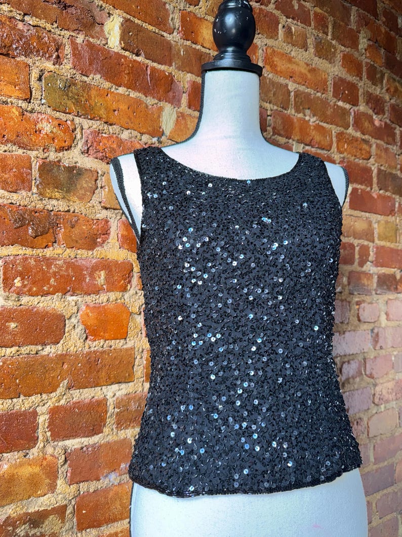 K&ouml;nnte beinhalten: Ein schwarzes Pailletten-Tanktop auf einer Schaufensterpuppe. Das &auml;rmellose Oberteil ist mit kleinen, gl&auml;nzenden Pailletten bedeckt. Es hat einen Rundhalsausschnitt und eine taillierte Silhouette. Der Hintergrund ist eine Backsteinmauer.