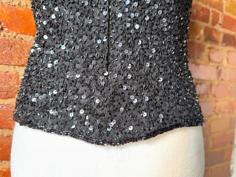K&ouml;nnte beinhalten: Schwarzes Pailletten-Top mit eng anliegender Silhouette. Das Kleidungsst&uuml;ck ist dicht mit kleinen, reflektierenden Pailletten bedeckt, wodurch ein schimmernder Effekt entsteht. Das Top wird auf einer wei&szlig;en Schaufensterpuppe mit einer Backsteinmauer im Hintergrund pr&auml;sentiert.