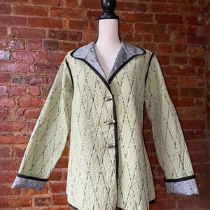 May include: A light green quilted jacket with a diamond pattern and black trim. The jacket has long sleeves, a collar, and three silver-tone toggle closures. The cuffs and collar lining feature a gray and black patterned fabric.