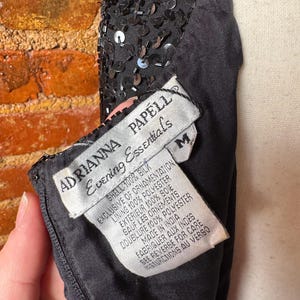 K&ouml;nnte beinhalten: Schwarzes Abendkleid mit Paillettenbesatz. Das Kleid hat ein Etikett mit der Aufschrift "ADRIANNA PAPELL Evening Essentials" und listet die Materialien Seide und Polyester auf. Das Kleid ist Gr&ouml;&szlig;e M.