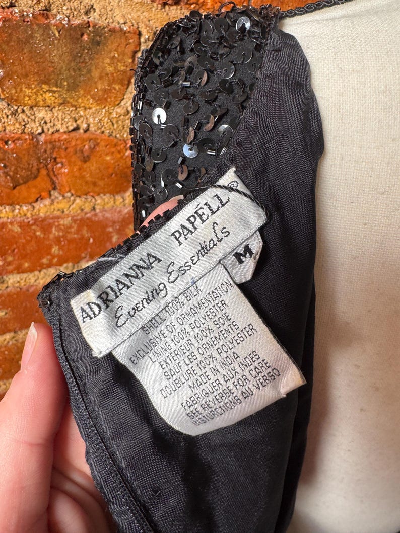 K&ouml;nnte beinhalten: Schwarzes Abendkleid mit Paillettenbesatz. Das Kleid hat ein Etikett mit der Aufschrift "ADRIANNA PAPELL Evening Essentials" und listet die Materialien Seide und Polyester auf. Das Kleid ist Gr&ouml;&szlig;e M.