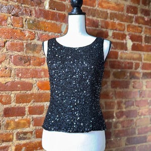K&ouml;nnte beinhalten: Ein schwarzes Pailletten-Tanktop auf einer Schaufensterpuppe. Das &auml;rmellose Oberteil hat einen Rundhalsausschnitt und ist mit kleinen, gl&auml;nzenden Pailletten bedeckt. Die Schaufensterpuppe steht vor einer Backsteinmauer.