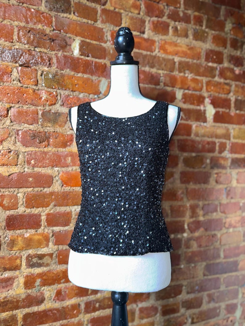 K&ouml;nnte beinhalten: Ein schwarzes Pailletten-Tanktop auf einer Schaufensterpuppe. Das &auml;rmellose Oberteil hat einen Rundhalsausschnitt und ist mit kleinen, gl&auml;nzenden Pailletten bedeckt. Die Schaufensterpuppe steht vor einer Backsteinmauer.