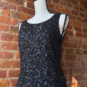 K&ouml;nnte beinhalten: Ein schwarzes Pailletten-Tanktop, das auf einer Schaufensterpuppe pr&auml;sentiert wird. Das &auml;rmellose Oberteil ist mit kleinen, gl&auml;nzenden Pailletten bedeckt. Der Ausschnitt ist rund und das Oberteil hat eine taillierte Silhouette. Der Hintergrund ist eine Backsteinmauer.