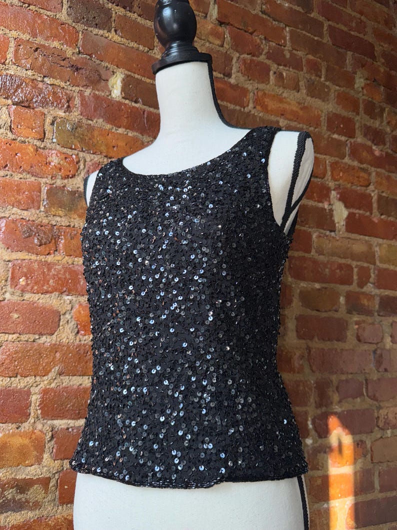 K&ouml;nnte beinhalten: Ein schwarzes Pailletten-Tanktop, das auf einer Schaufensterpuppe pr&auml;sentiert wird. Das &auml;rmellose Oberteil ist mit kleinen, gl&auml;nzenden Pailletten bedeckt. Der Ausschnitt ist rund und das Oberteil hat eine taillierte Silhouette. Der Hintergrund ist eine Backsteinmauer.