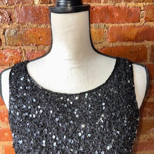 K&ouml;nnte beinhalten: Schwarzes &auml;rmelloses Kleid mit Pailletten. Das Kleid wird auf einer Schaufensterpuppe vor einer Backsteinmauer pr&auml;sentiert. Die Pailletten sind eng aneinandergereiht und erzeugen einen schimmernden Effekt.