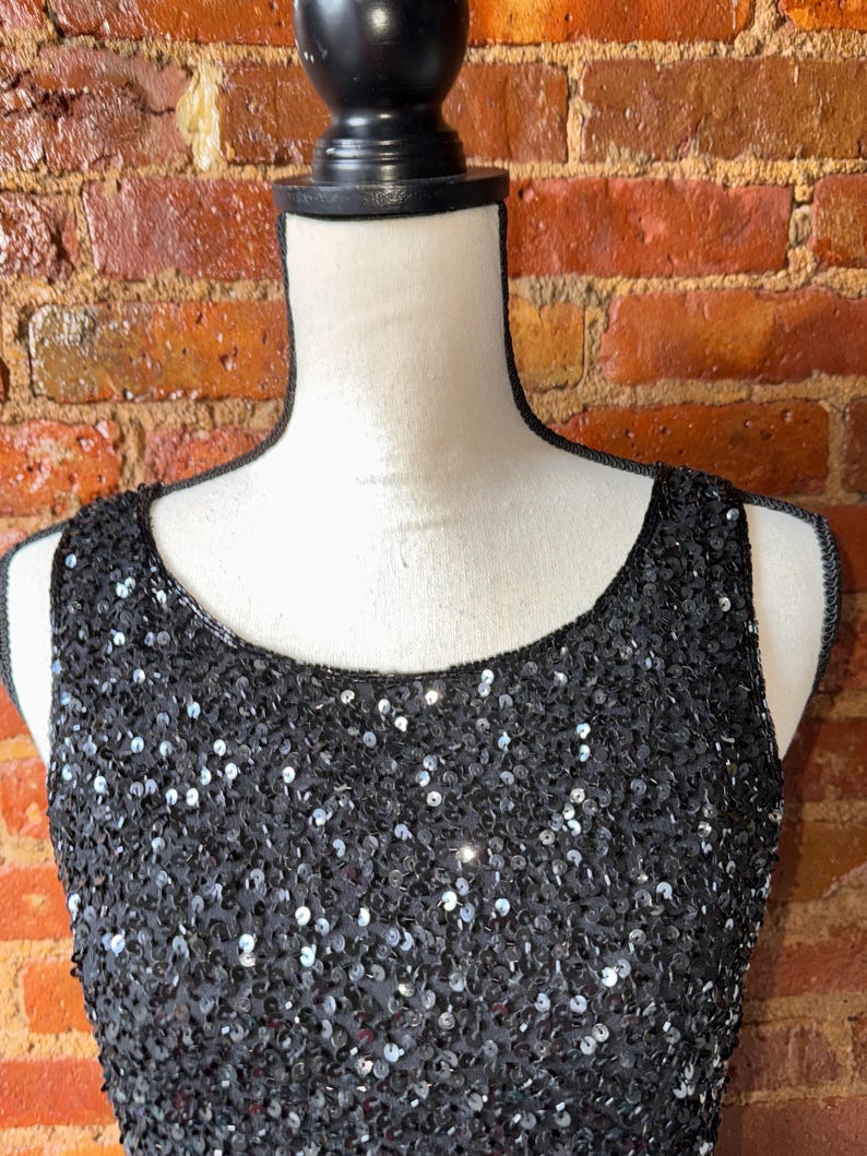 K&ouml;nnte beinhalten: Schwarzes &auml;rmelloses Kleid mit Pailletten. Das Kleid wird auf einer Schaufensterpuppe vor einer Backsteinmauer pr&auml;sentiert. Die Pailletten sind eng aneinandergereiht und erzeugen einen schimmernden Effekt.
