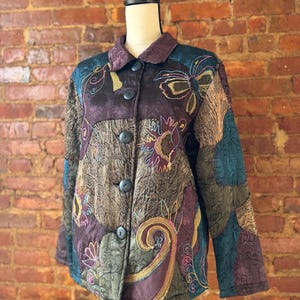 May include: A quilted jacket with a patchwork design in shades of brown, teal, and purple. The jacket features embroidered floral and paisley patterns, with large black buttons down the front. The collar is a darker shade of purple.
