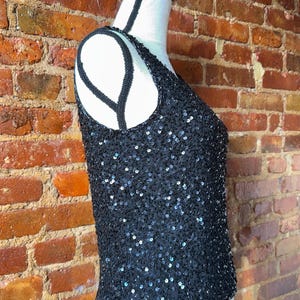 K&ouml;nnte beinhalten: Ein schwarzes Pailletten-Tanktop auf einer Schaufensterpuppe. Das Oberteil ist mit kleinen, gl&auml;nzenden Pailletten bedeckt und hat einen Rundhalsausschnitt. Die Tr&auml;ger und der Ausschnitt sind mit schwarzen Perlen verziert. Der Hintergrund ist eine Backsteinmauer.
