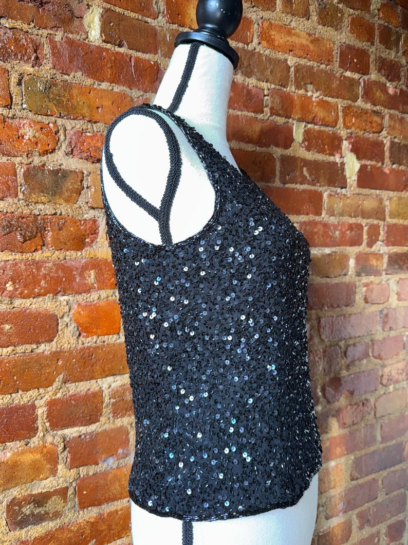 K&ouml;nnte beinhalten: Ein schwarzes Pailletten-Tanktop auf einer Schaufensterpuppe. Das Oberteil ist mit kleinen, gl&auml;nzenden Pailletten bedeckt und hat einen Rundhalsausschnitt. Die Tr&auml;ger und der Ausschnitt sind mit schwarzen Perlen verziert. Der Hintergrund ist eine Backsteinmauer.