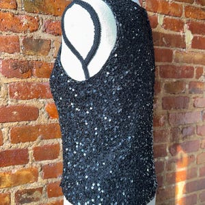 K&ouml;nnte beinhalten: Schwarzes Pailletten-Tanktop mit Rundhalsausschnitt und &auml;rmellosem Design. Das Oberteil ist mit kleinen, gl&auml;nzenden Pailletten bedeckt, die einen schimmernden Effekt erzeugen. Der Halsausschnitt und die Armausschnitte sind mit einer schwarzen Kordel versehen. Auf einer wei&szlig;en Schaufensterpuppe pr&auml;sentiert.