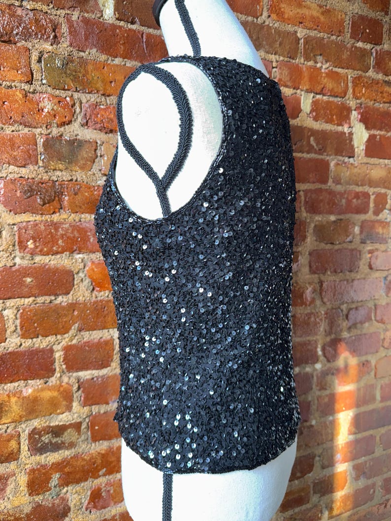 K&ouml;nnte beinhalten: Schwarzes Pailletten-Tanktop mit Rundhalsausschnitt und &auml;rmellosem Design. Das Oberteil ist mit kleinen, gl&auml;nzenden Pailletten bedeckt, die einen schimmernden Effekt erzeugen. Der Halsausschnitt und die Armausschnitte sind mit einer schwarzen Kordel versehen. Auf einer wei&szlig;en Schaufensterpuppe pr&auml;sentiert.