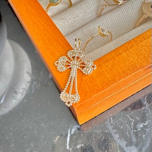 Può includere: Un ciondolo a forma di croce color oro con un design dettagliato e ornato. La croce è in metallo e presenta un motivo delicato e intricato.