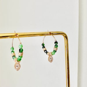 Peut inclure: Boucles d'oreilles créoles dorées ornées de perles vertes et dorées. Chaque boucle d'oreille est ornée d'un charm en forme de goutte avec des cristaux transparents. Les boucles d'oreilles sont présentées sur un présentoir à bijoux doré.